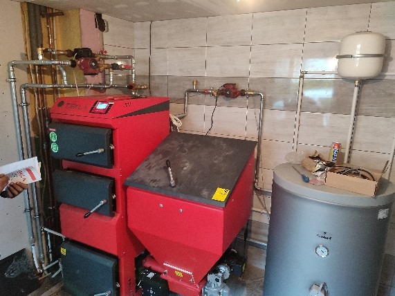 Czerwony piec centralnego ogrzewania marki Tekla z podajnikiem, zbiornik na wodę z manometrem, rury instalacji CO i zawory w kotłowni wyłożonej płytkami.