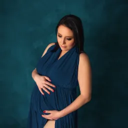 Kobieta w zaawansowanej ciąży, ubrana w długą, granatową suknię z rozcięciem, pozuje na tle teksturowanej, turkusowej ściany, dłonie spoczywają na brzuchu, podkreślając jego kształt.