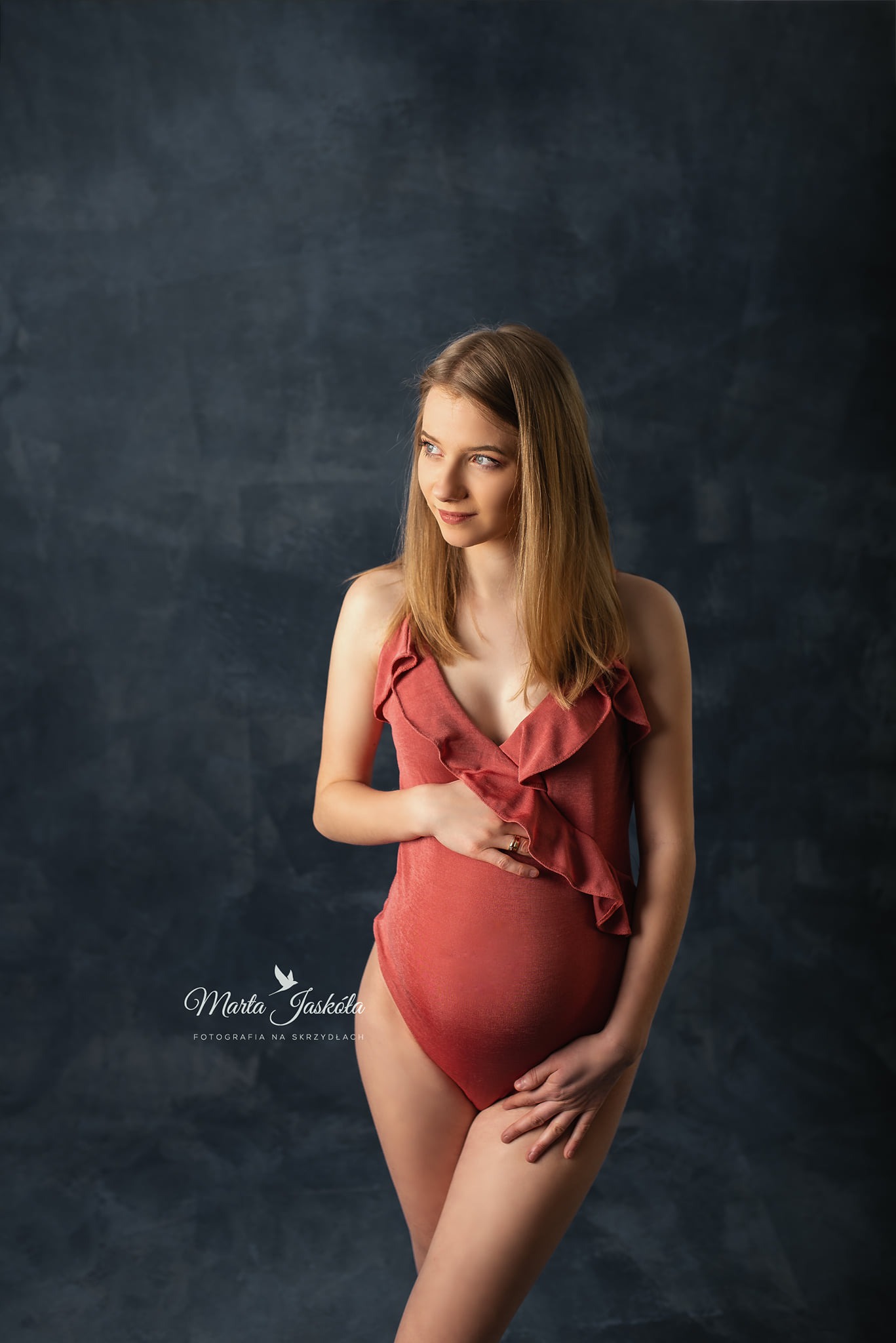 Portret kobiety w ciąży, w koralowym kostiumie z falbankami, dłonie na brzuchu, złoty pierścionek, ciemne tło z efektem malarskim.