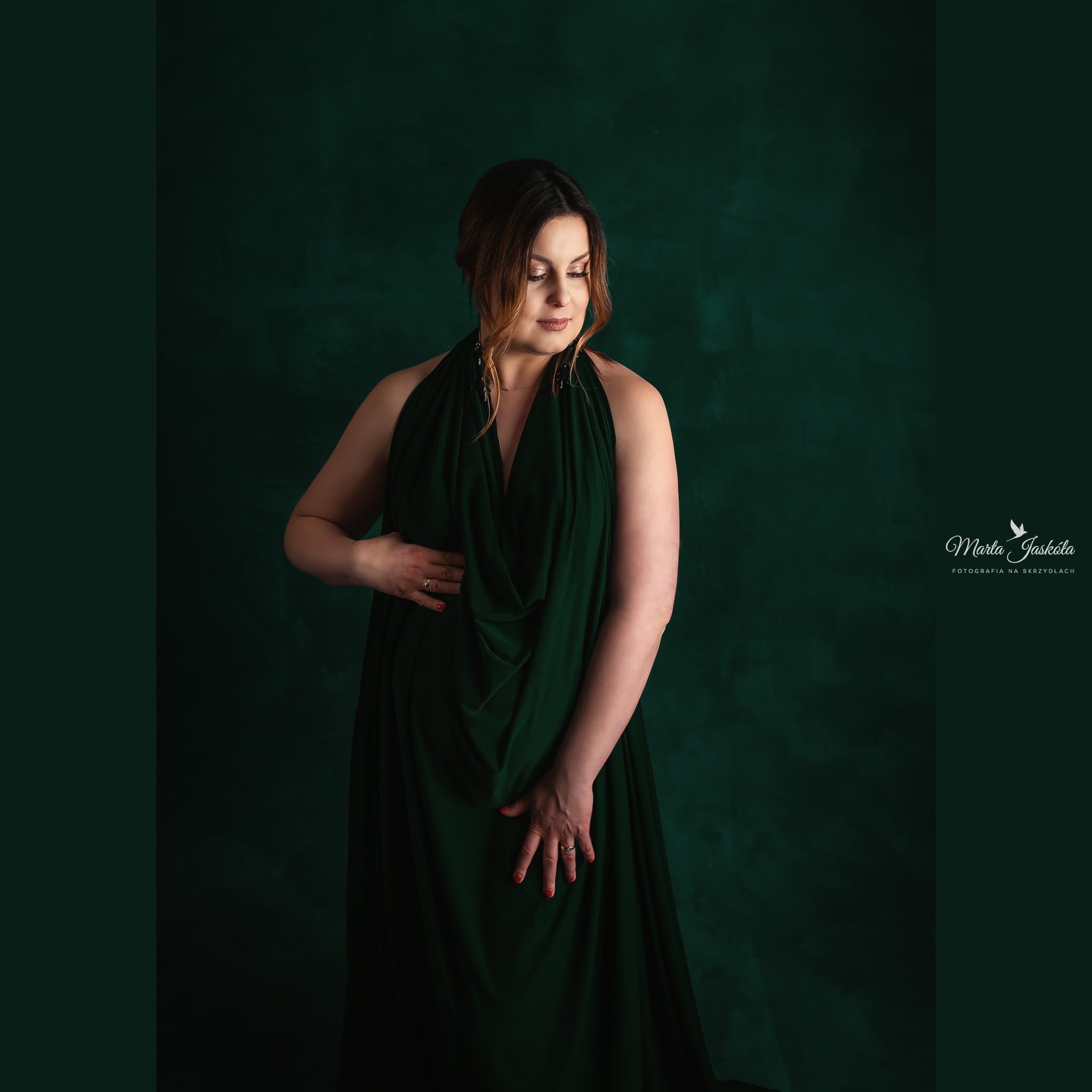 Portret kobiety w ciąży, w zielonej drapowanej sukni na tle ciemnozielonego tła, dłonie spoczywają na brzuchu, delikatny makijaż.