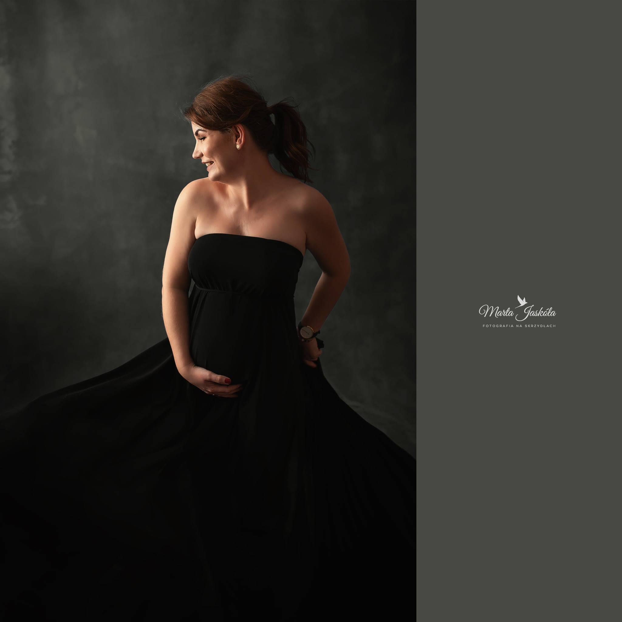 Uśmiechnięta kobieta w ciąży, ubrana w czarną sukienkę bez ramiączek, trzyma ręce na brzuchu, profil na szarym tle, widoczny zegarek na ręce.