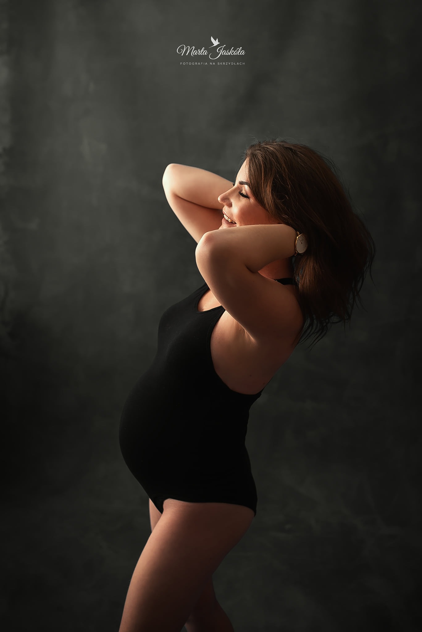 Kobieta w czarnym body, z włosami upiętymi do góry, pozuje z uśmiechem, eksponując ciążowy brzuszek na tle ciemnego, teksturowanego tła. Na nadgarstku widoczny złoty zegarek.