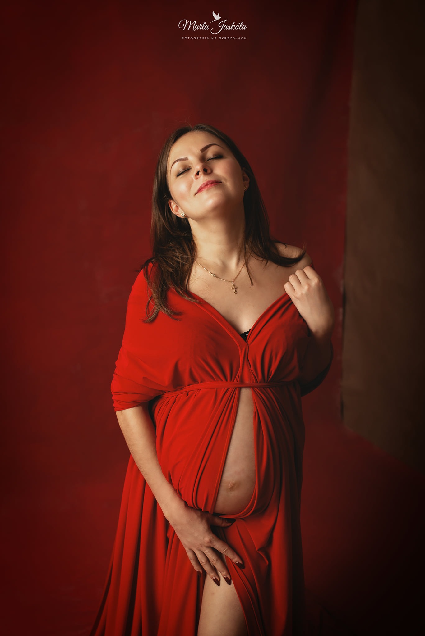 Kobieta w czerwonej sukni, z zamkniętymi oczami, delikatnie dotyka brzucha na tle bordowej ściany.