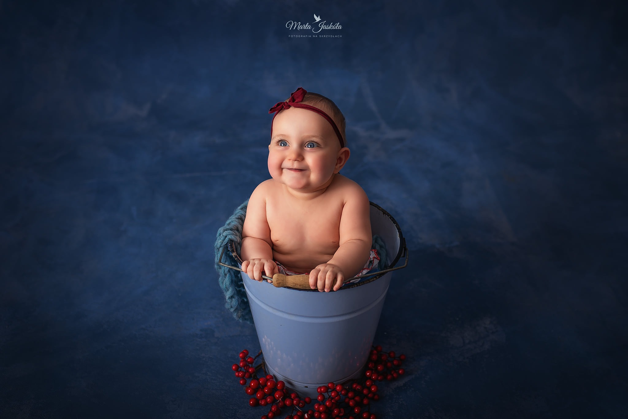 Uśmiechnięta niemowlęca dziewczynka z czerwoną kokardą na głowie siedzi w jasnoniebieskim wiaderku na tle ciemnoniebieskiego tła z dekoracją z czerwonych jagód.