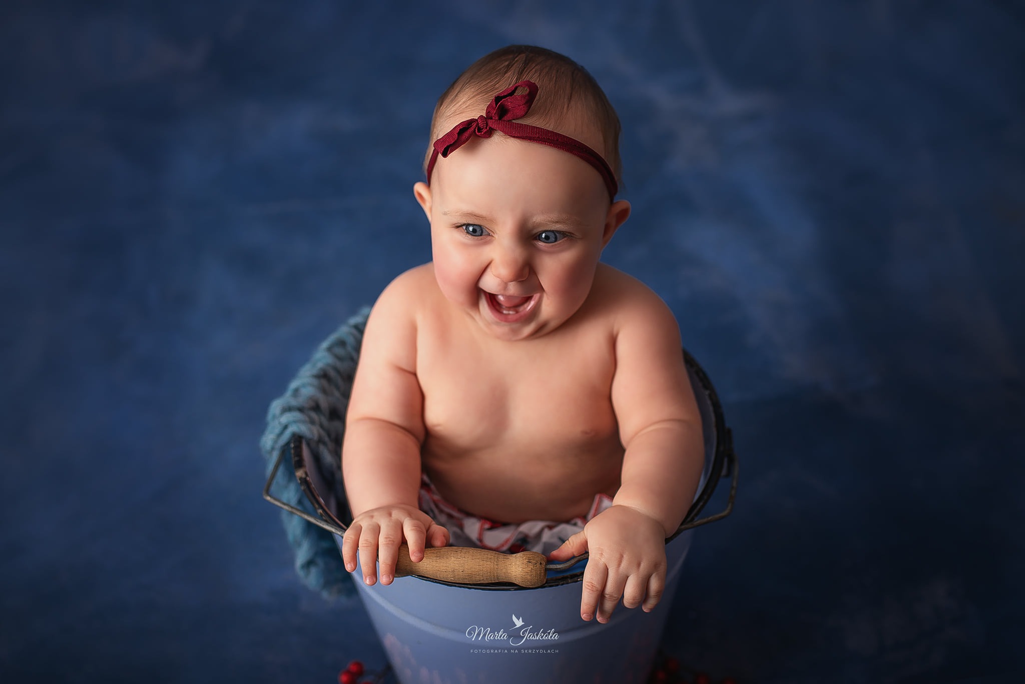 Uśmiechnięta niemowlęca dziewczynka z czerwoną opaską na głowie, siedząca w niebieskim wiaderku z drewnianą rączką na tle ciemnoniebieskiego tła.