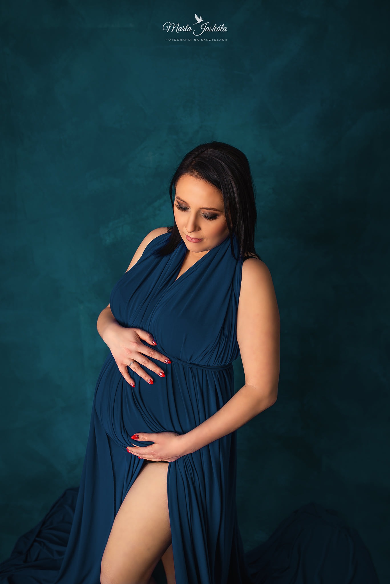Kobieta w zaawansowanej ciąży, ubrana w długą, granatową suknię z rozcięciem, pozuje na tle teksturowanej, turkusowej ściany, dłonie spoczywają na brzuchu, podkreślając jego kształt.
