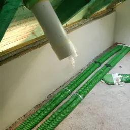 Fragment instalacji rekuperacji z zielonymi rurami karbowanymi, białym pionowym kanałem i elementami mocującymi pod drewnianą konstrukcją dachu.