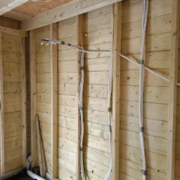 Wnętrze szkieletowej konstrukcji drewnianej z poprowadzonymi rurami wentylacyjnymi, tymczasowo przymocowanymi taśmą do słupków.