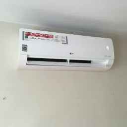 Biała jednostka wewnętrzna klimatyzatora LG Dual Inverter zamontowana na jasnej ścianie, widoczny wyświetlacz temperatury i naklejka informująca o oszczędności energii.