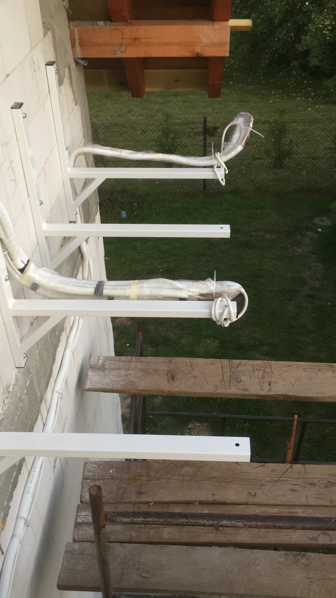 Białe wsporniki montażowe na ścianie z białej cegły, przygotowane do instalacji rur, widoczne tymczasowe rusztowanie drewniane i trawnik w tle.