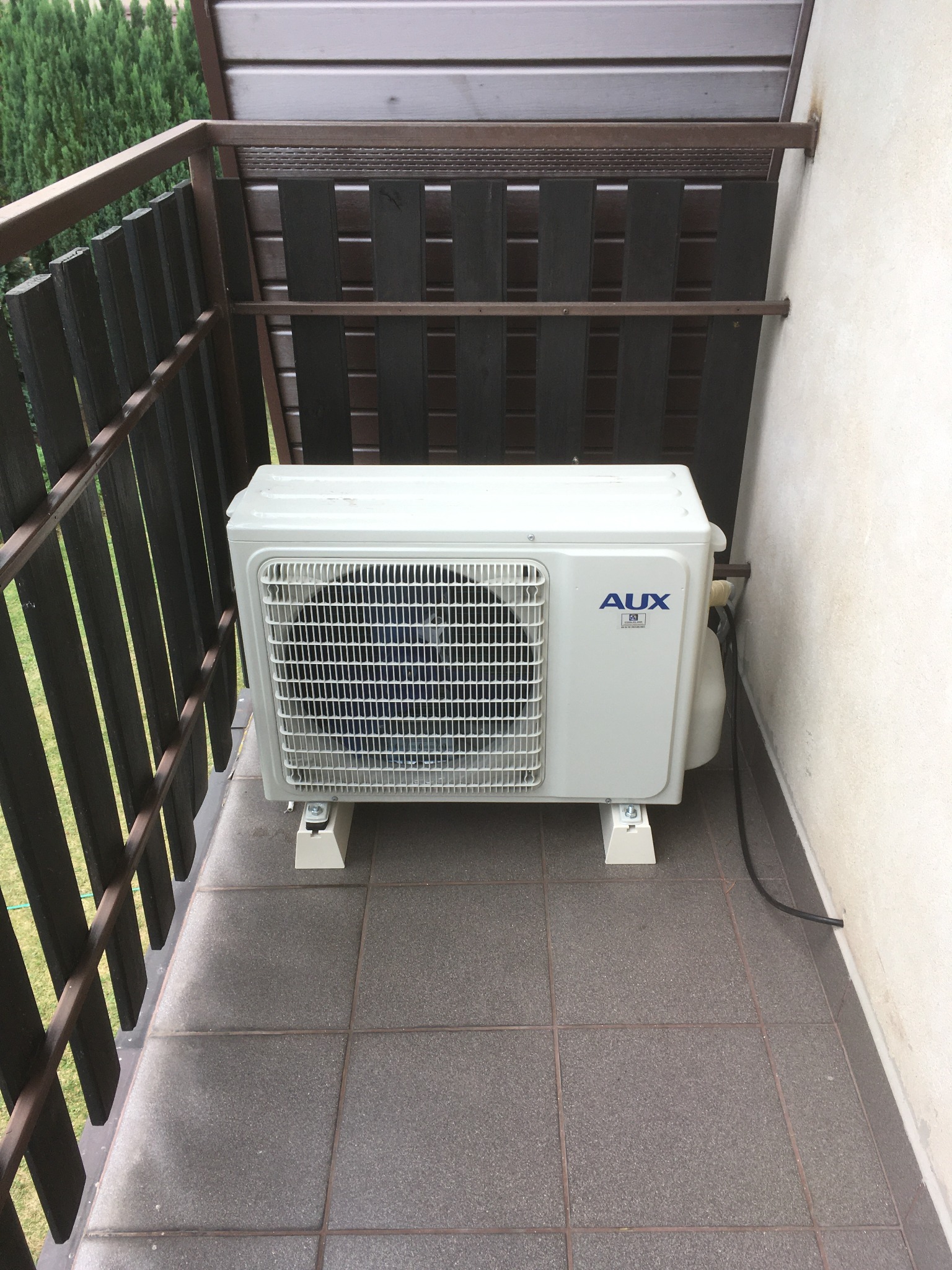 Biały klimatyzator AUX zamontowany na balkonie z brązową balustradą, widoczny kabel zasilający i fragment ściany budynku.