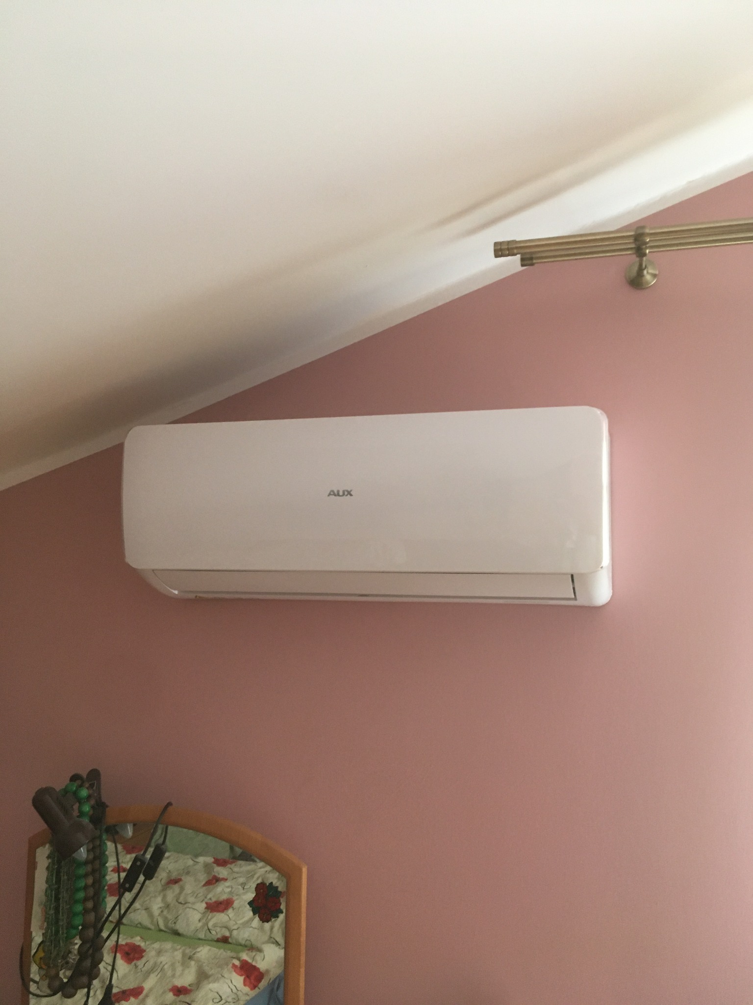 Biała jednostka klimatyzacji AUX zamontowana na różowej ścianie w pokoju z ukośnym sufitem, widoczna część lustra z odbiciem pościeli z czerwonymi makami.