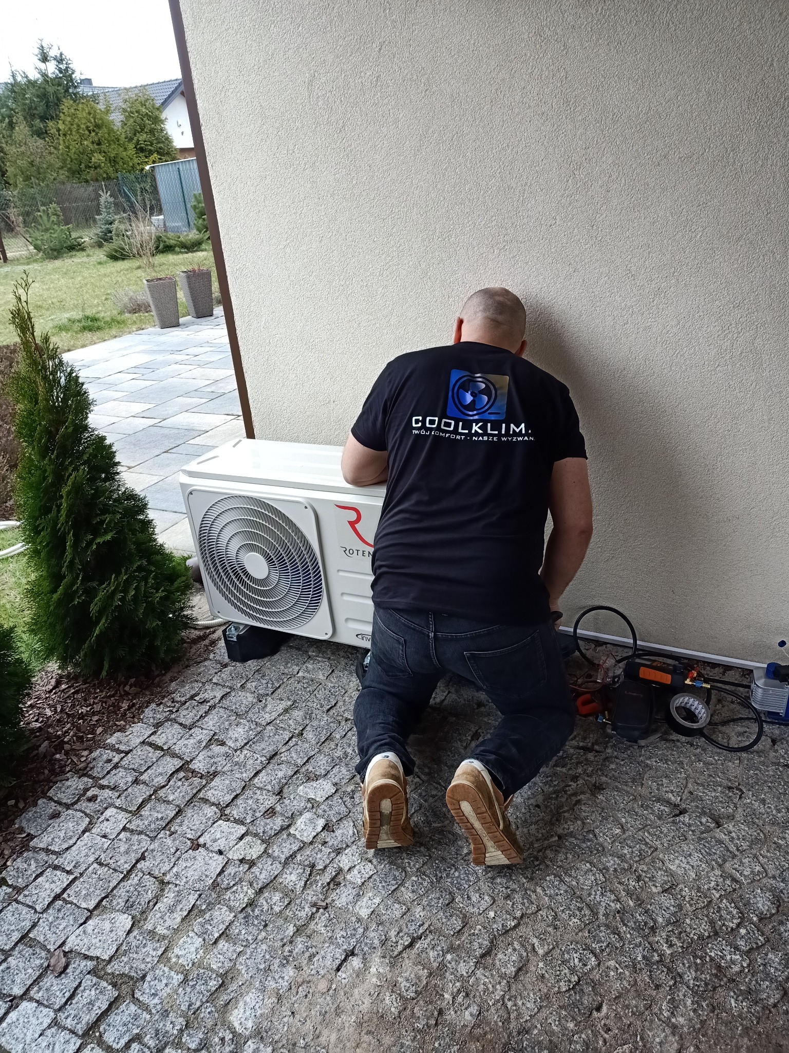 Instalator w czarnej koszulce z logo firmy COOLKLIM, klęczy na kostce brukowej obok zewnętrznej jednostki klimatyzacji Roten, podłączając przewody i urządzenia serwisowe do instalacji.