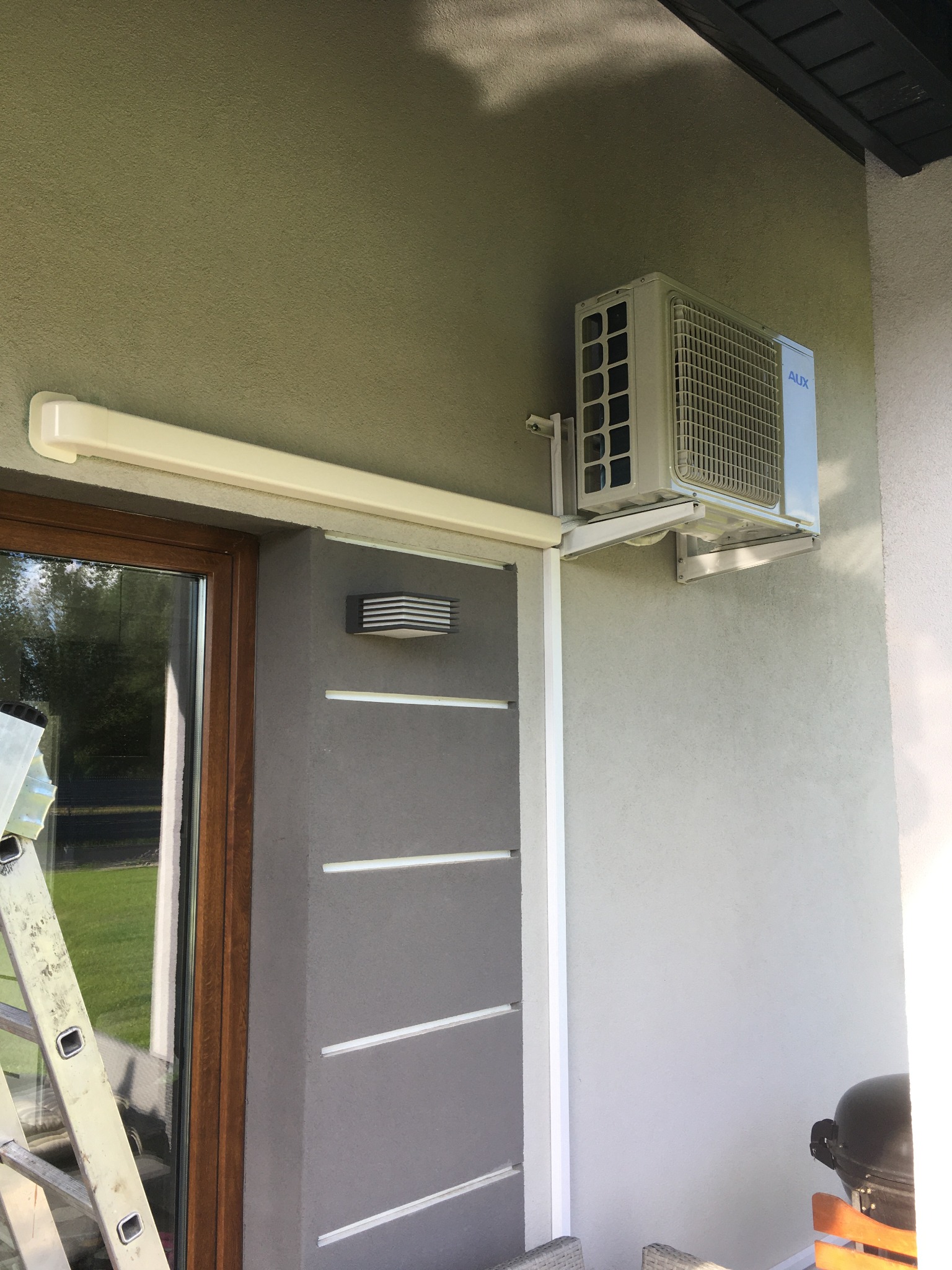 Zewnętrzna jednostka klimatyzatora marki AUX zamontowana na ścianie domu z widoczną drabiną opartą o okno.