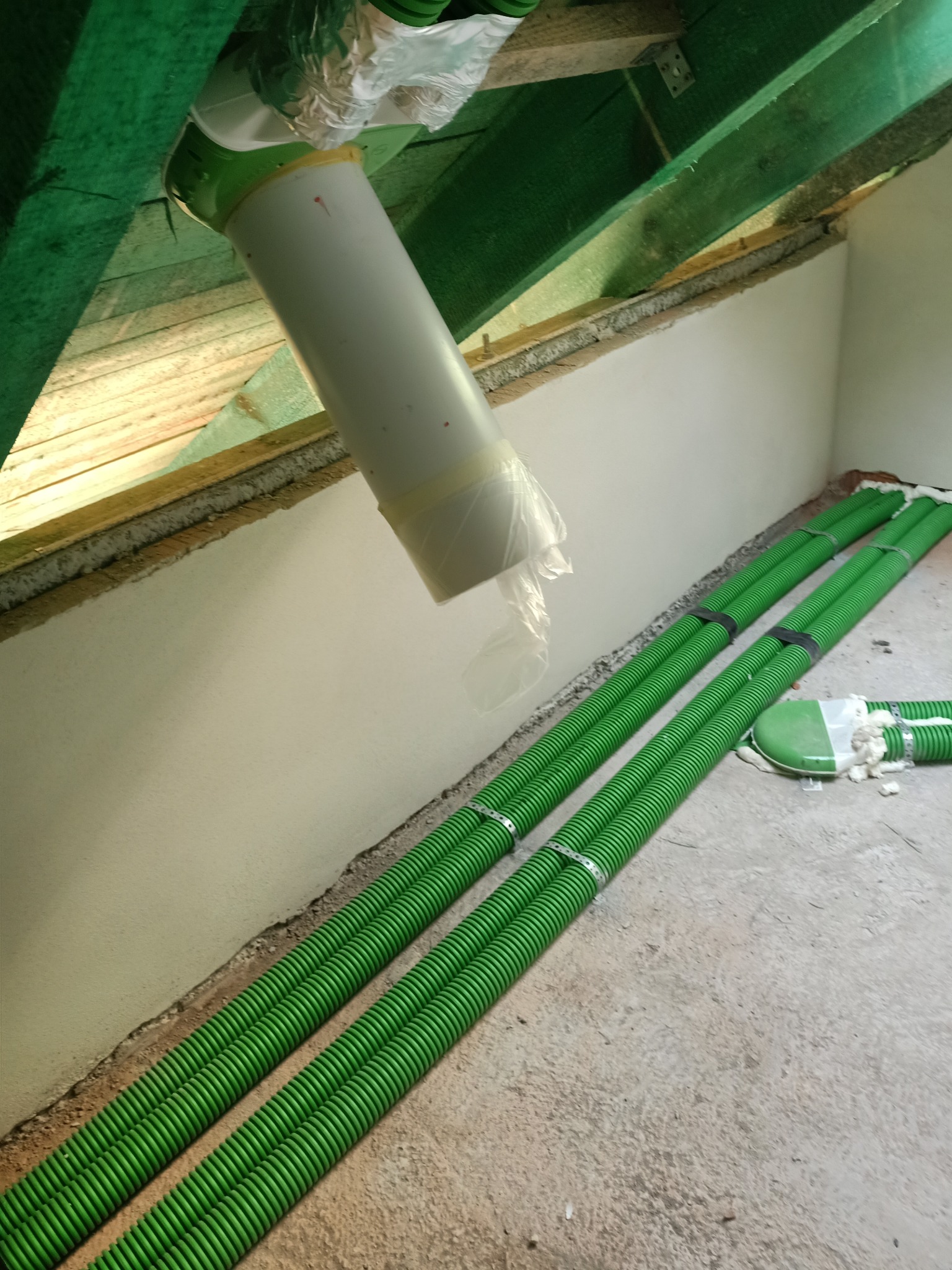 Fragment instalacji rekuperacji z zielonymi rurami karbowanymi, białym pionowym kanałem i elementami mocującymi pod drewnianą konstrukcją dachu.