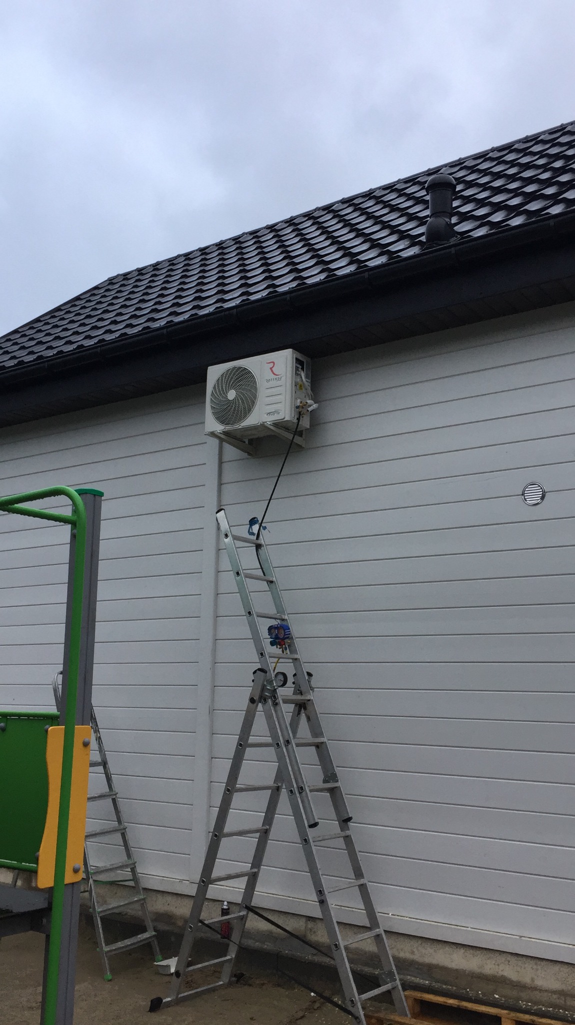Zewnętrzna jednostka klimatyzacji zamontowana na elewacji domu, widoczna drabina i narzędzia serwisowe, obok plac zabaw dla dzieci.