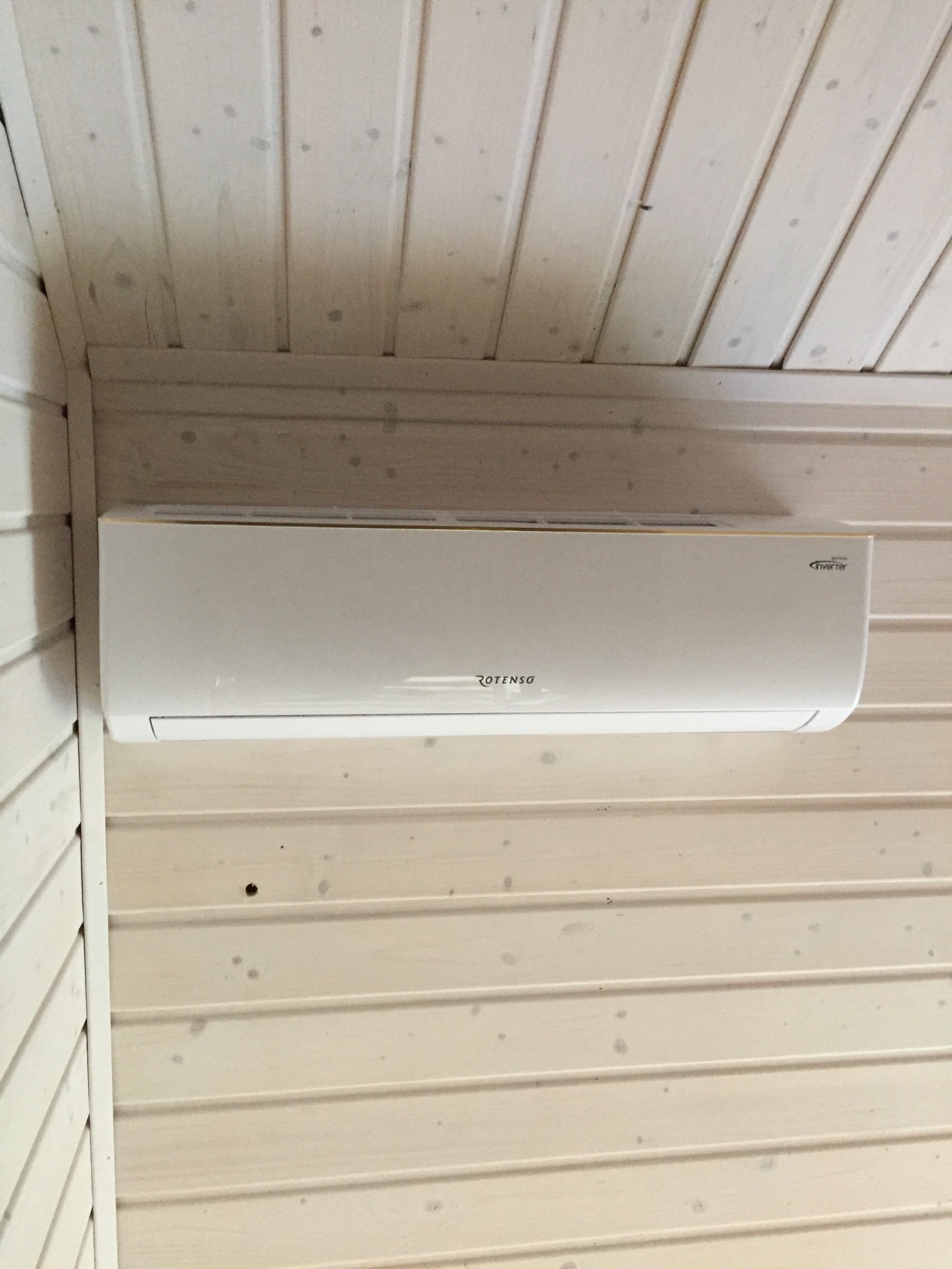 Biała klimatyzacja Rotenso Inverter zamontowana na jasnej, drewnianej ścianie z boazerią, widoczne sęki i naturalna struktura drewna.
