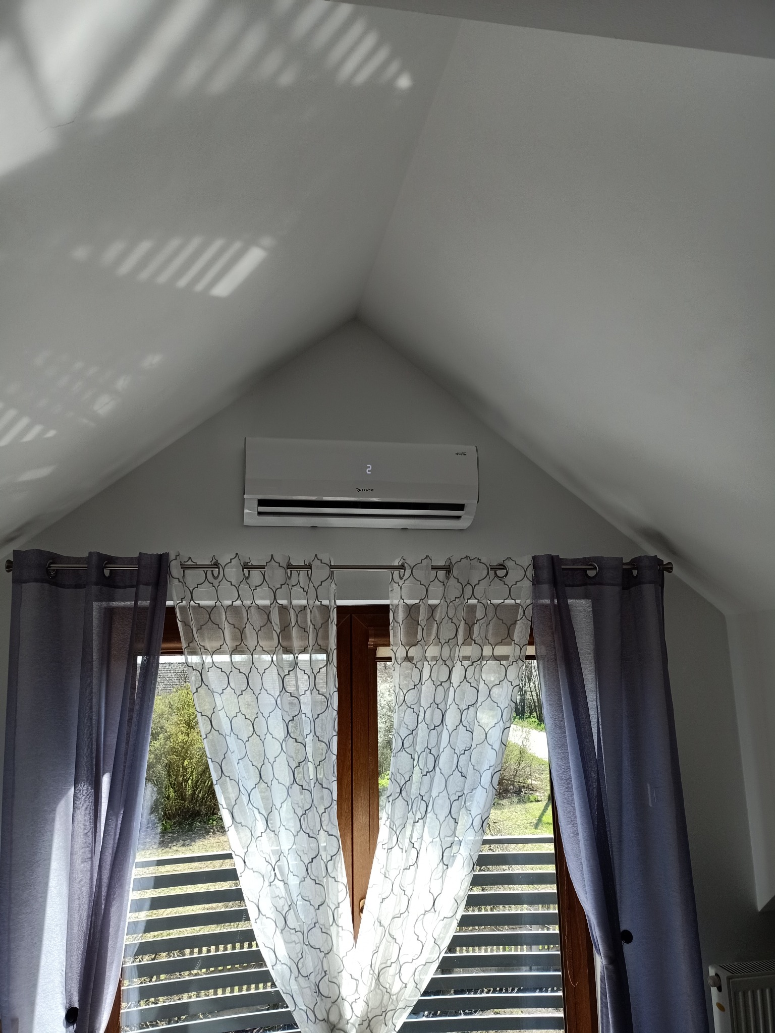 Biały klimatyzator marki Rotenso zamontowany na ścianie pod skosem, widoczny nad oknem z zasłonami w pokoju na poddaszu.