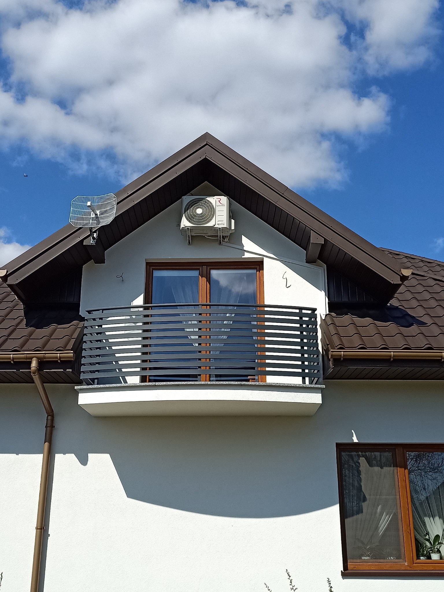 Biały budynek z brązowym dachem, balkonem i zamontowaną jednostką zewnętrzną klimatyzacji pod szczytem dachu, widoczna antena na dachu.