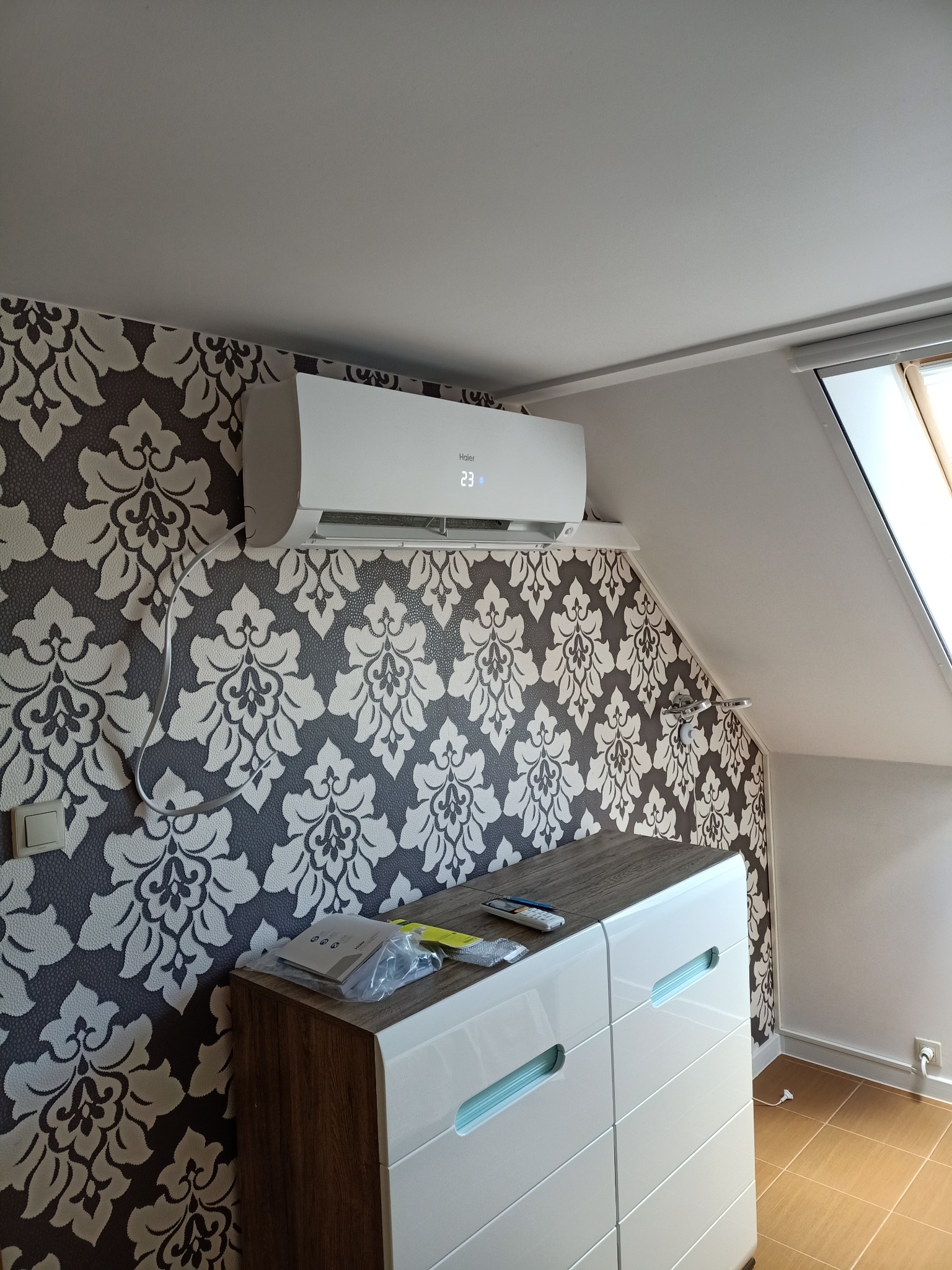 Biały klimatyzator Haier zamontowany na ścianie z wzorzystą tapetą w pokoju na poddaszu, widoczne akcesoria montażowe na komodzie pod spodem.