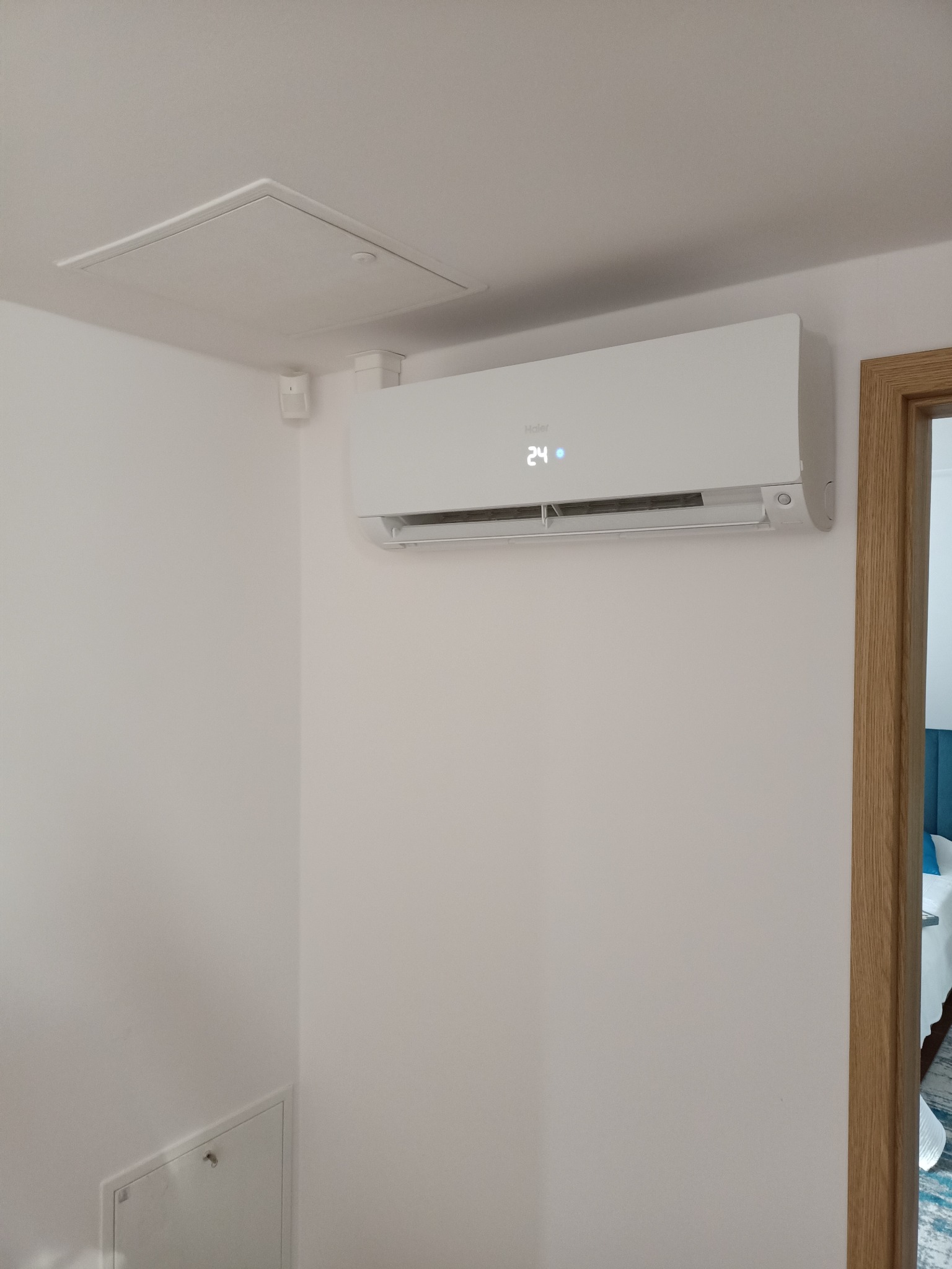 Biały klimatyzator ścienny Haier z wyświetlaczem temperatury 24°C, zamontowany w rogu pokoju, widoczna klapa rewizyjna na suficie i fragment drewnianej ościeżnicy drzwiowej.