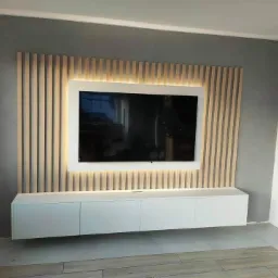 Nowoczesna ściana telewizyjna z podświetlonym panelem drewnianym i białą szafką RTV zawieszoną pod spodem, na tle szarej ściany.