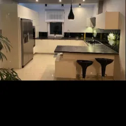 Nowoczesna kuchnia w odcieniach bieli i szarości, z czarnym blatem i barkiem z dwoma hokerami, oświetlona ledami i lampą wiszącą.