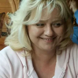 Portret uśmiechniętej kobiety z blond włosami w różowej bluzie na tle drewnianej ściany z kwiatowym ornamentem.