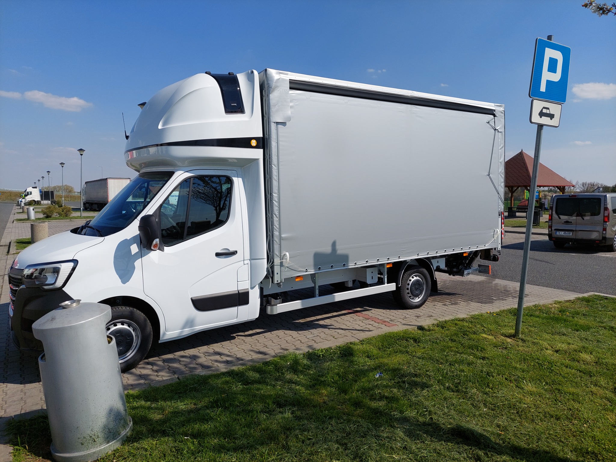 Biały samochód dostawczy z plandeką zaparkowany na parkingu przy drodze, obok kosza na śmieci i znaku drogowego 'parking'.