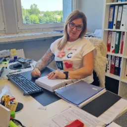 Kobieta w okularach z tatuażem na ramieniu, siedząca przy biurku z komputerem, segregatorami i kalkulatorem, wypełniająca dokumenty długopisem.