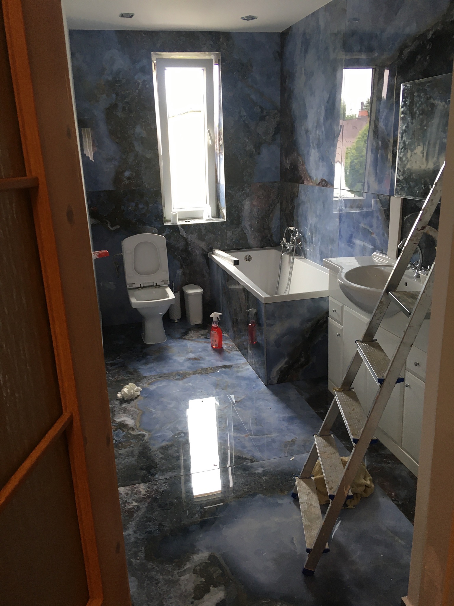 Wnętrze łazienki w trakcie remontu z marmurowymi płytkami w odcieniach niebieskiego, widoczna drabina, toaleta, wanna, umywalka z szafką oraz butelka środka czyszczącego.
