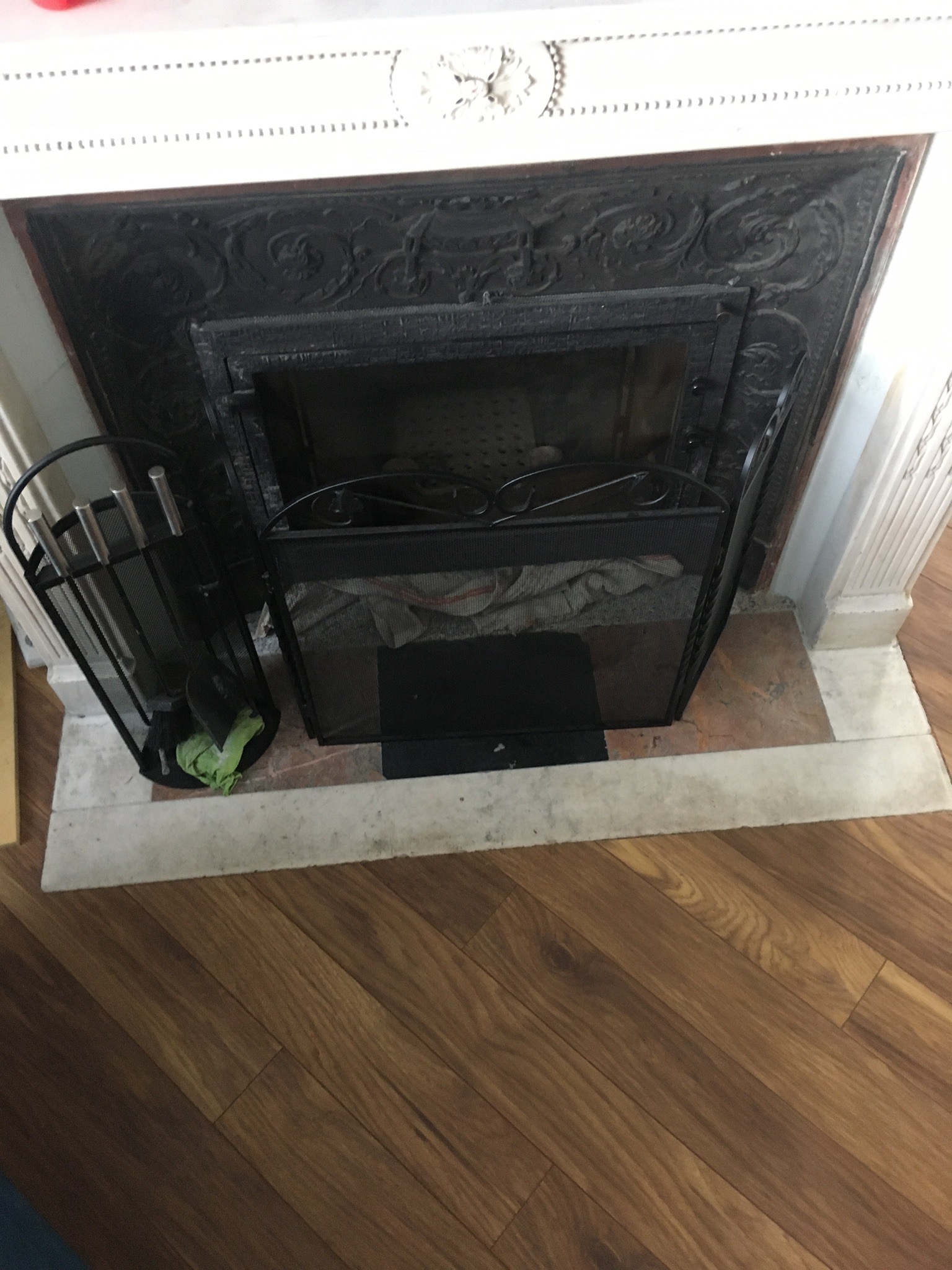 Ozdobny kominek z czarnym wkładem, metalowym ekranem ochronnym i zestawem pogrzebaczy, na tle marmurowego obramowania i drewnianej podłogi.