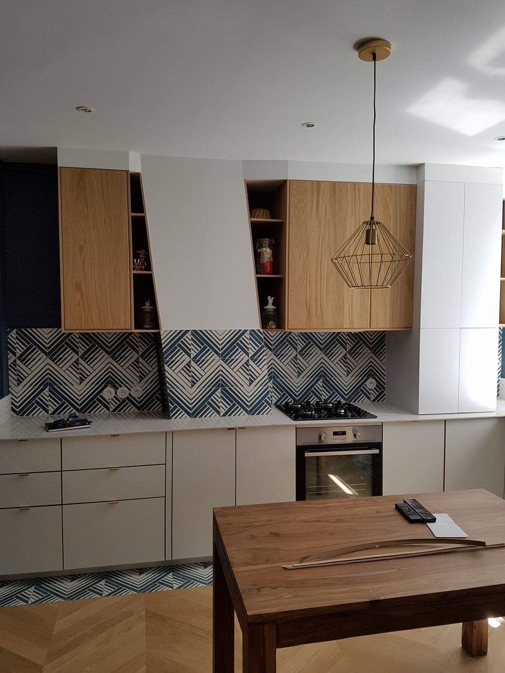Nowoczesna kuchnia z drewnianymi frontami szafek górnych, wzorzystą mozaiką na ścianie nad blatem, drewnianym stołem i złotą lampą wiszącą o geometrycznym kształcie
