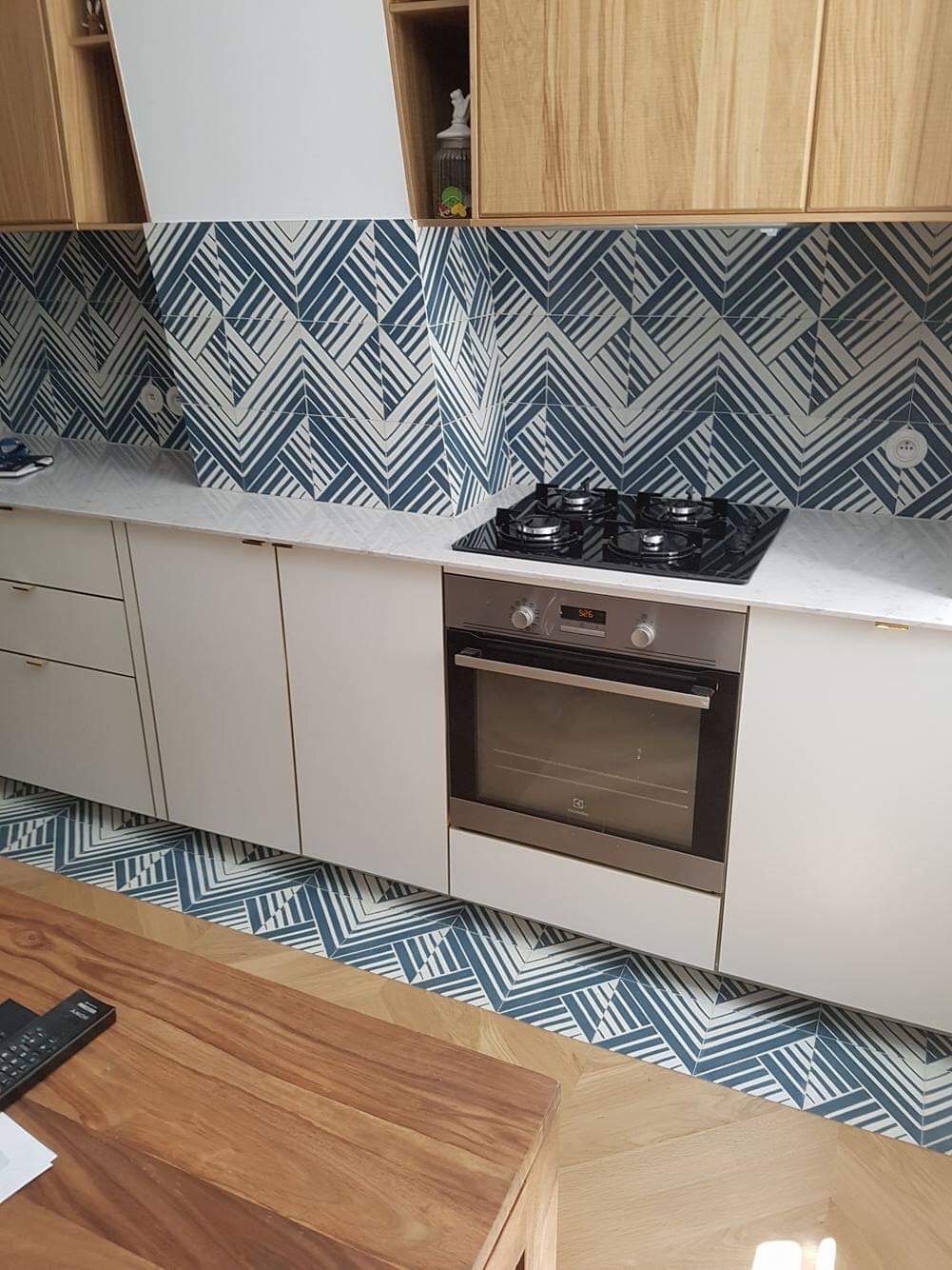 Kuchnia z białymi szafkami, drewnianymi górnymi szafkami, blatem z efektem marmuru oraz niebiesko-białymi płytkami z geometrycznym wzorem na ścianie i podłodze przy szafkach.