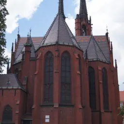 Ceglana bryła kościoła gotyckiego z wysokimi wieżami i spadzistym dachem pokrytym czerwoną dachówką, widoczna z poziomu gruntu, otoczona zielenią.