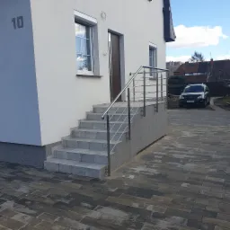 Zewnętrzne schody wejściowe do domu z jasną elewacją, zabezpieczone balustradą ze stali nierdzewnej o minimalistycznym designie, widok z poziomu kostki brukowej.