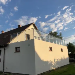 Biały dom z płaskim dachem i balkonem zabezpieczonym szklaną balustradą z nierdzewnymi słupkami, na tle błękitnego nieba z chmurami.