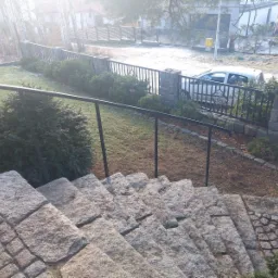 Granitowe schody zewnętrzne z czarną metalową balustradą, prowadzące do ogrodu z widocznym ogrodzeniem z kamiennymi słupkami i metalowymi przęsłami oraz zaparkowanym samochodem w tle.