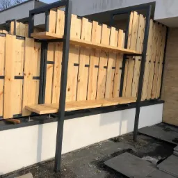 Drewniano-metalowa konstrukcja z półkami, tworząca ażurową przegrodę i element dekoracyjny na tarasie, widok w trakcie montażu, z widocznymi płytkami podłogowymi i narzędziami.