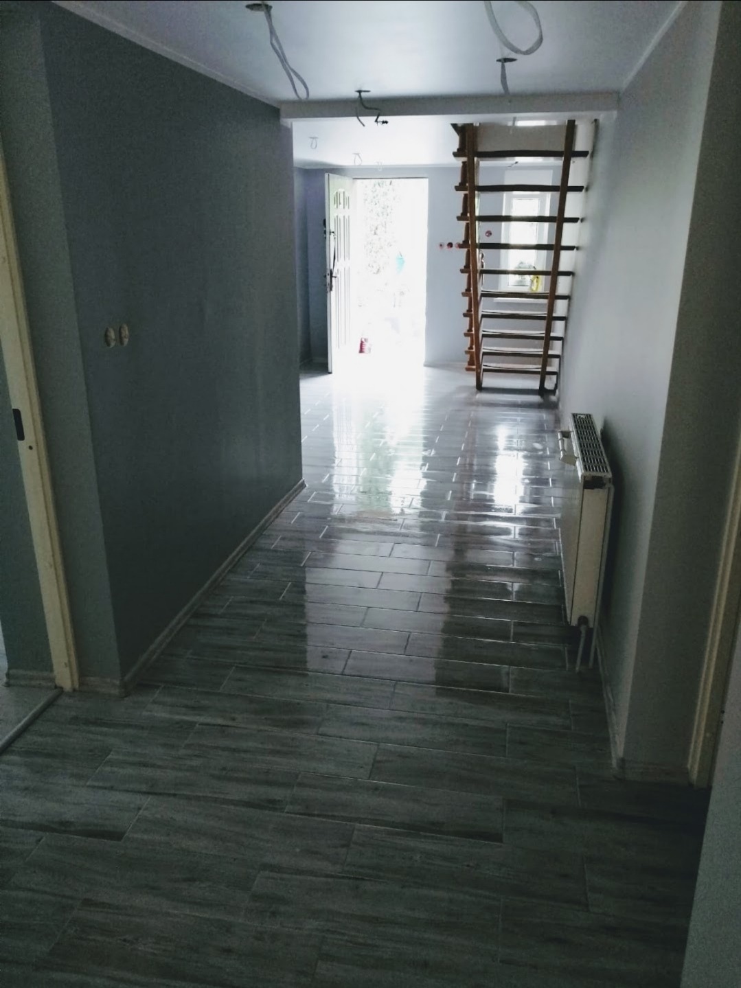 Korytarz po remoncie w Zgorzelcu: świeżo pomalowane szare ściany, błyszcząca podłoga z płytek imitujących drewno, widoczne kable wystające z sufitu i drewniana drabina oparta o ścianę.