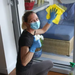 Kobieta w maseczce i niebieskich rękawiczkach czyści okno żółtą ściereczką, kucając na balkonie z rattanowymi meblami i roślinami doniczkowymi, kciuk w górę.