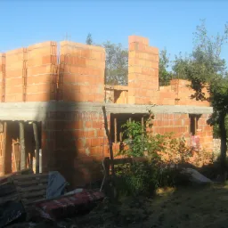 Surowy stan budynku z czerwonej cegły kratówki, widoczne zbrojenia pionowe, częściowo zabetonowany wieniec, paleta i gruz budowlany na pierwszym planie.