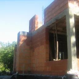 Wznoszenie ścian parterowego budynku z czerwonej cegły, widoczne drewniane podpory stropu, zbrojenie i fragment osoby w tle.