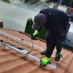 Instalator z neonowo-zielonymi rękawicami, w czarnym ubraniu i czapce z daszkiem, stojący na dachu z dachówki, przycina szlifierką kątową aluminiową konstrukcję pod panele fotowoltaiczne; w tle dom...