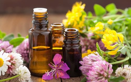 Trzy brązowe butelki z olejkami eterycznymi otoczone różnorodnymi kwiatami polnymi na drewnianym tle.
