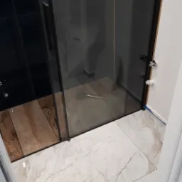 Nowoczesna kabina prysznicowa z czarnymi ramami i drzwiami przesuwnymi, widok na drewnopodobną podłogę wewnątrz kabiny i marmurowe płytki na zewnątrz, z widocznymi elementami instalacji wodnej...