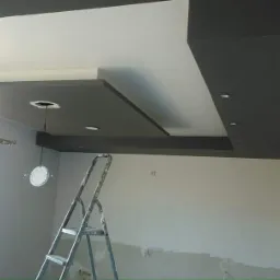 Dwupoziomowy sufit podwieszany z szarego karton gipsu z oświetleniem punktowym, widoczna drabina i przygotowane ściany do malowania.