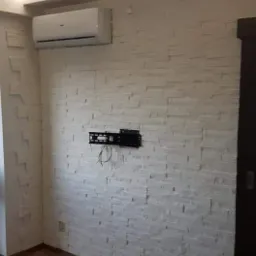 Ściana wykończona białą, dekoracyjną cegłą, z zamontowanym uchwytem do telewizora i klimatyzatorem, drewniana podłoga, widoczny fragment drzwi