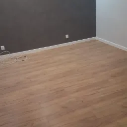 Pokój po remoncie z panelami podłogowymi w odcieniach brązu, szara ściana z dwoma kinkietami i biała ściana, widoczne gniazdka elektryczne i białe listwy przypodłogowe.