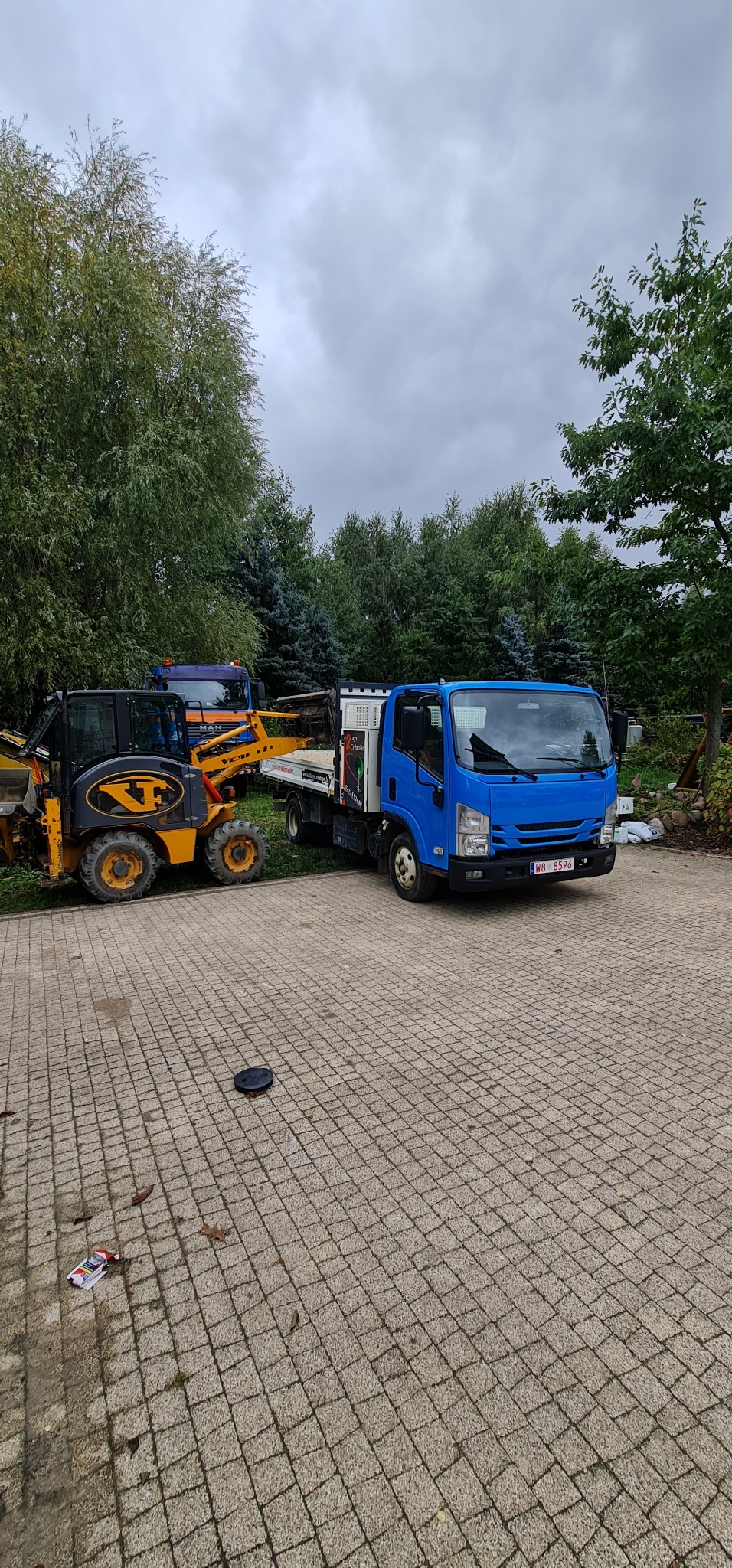 Niebieska ciężarówka z otwartą skrzynią ładunkową i żółta mini-ładowarka na kostce brukowej, w tle drzewa.